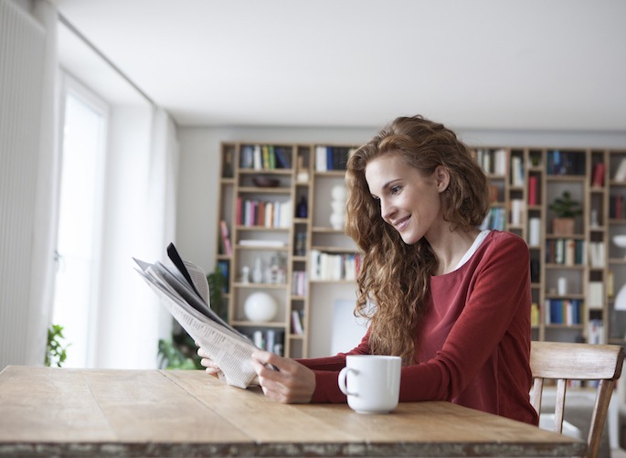 Mulher sorridente em casa, sentada numa mesa de madeira com uma chávena a ler um jornal
